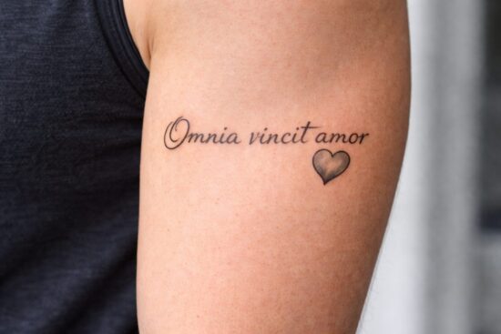 Idee tatuaggi amore latino: scritta in corsivo elegante 'Omnia vincit amor' (L'amore vince su tutto) con un cuore piccolo sul braccio