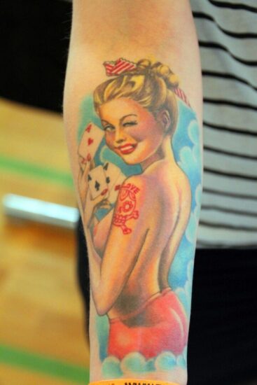 Tatuaggio a colori sull'avambraccio raffigurante una ragazza pin-up bionda in stile retrò che fa l'occhiolino e tiene in mano delle carte da gioco.